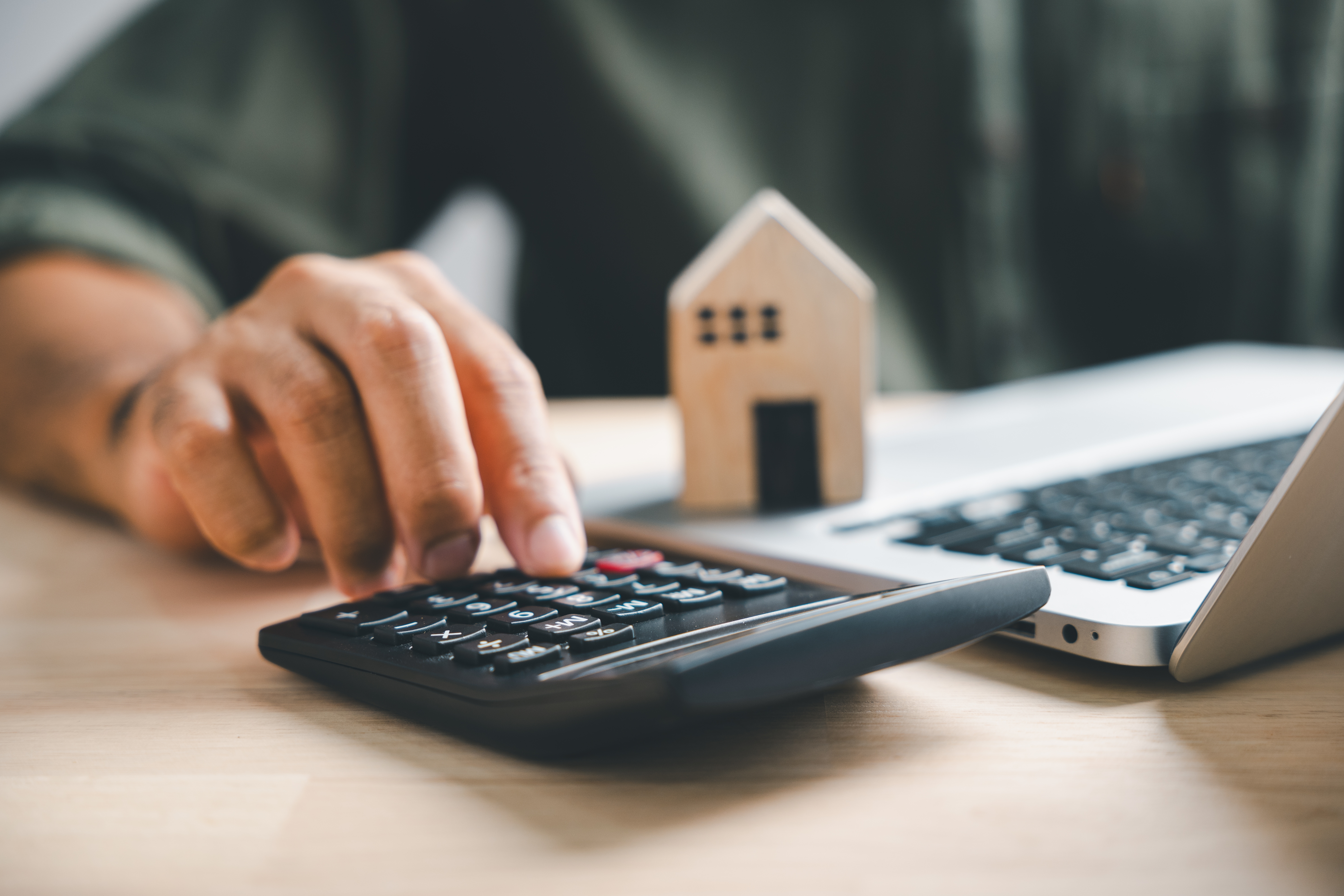 Hand typing on a calculator with small wooden house and laptop next to it