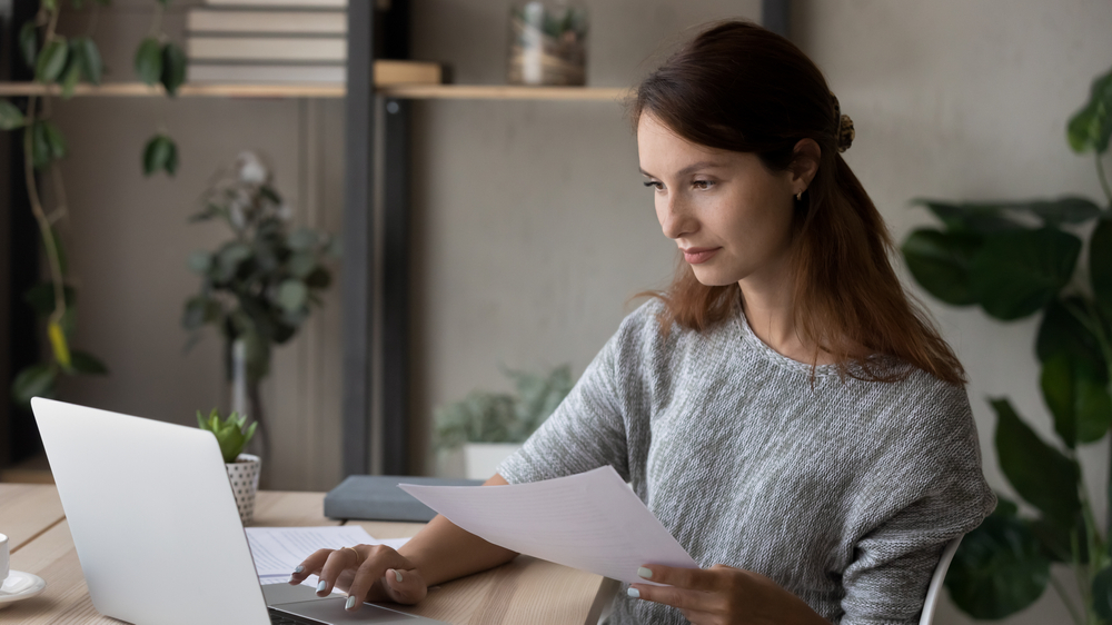 Young woman on laptop holding paper