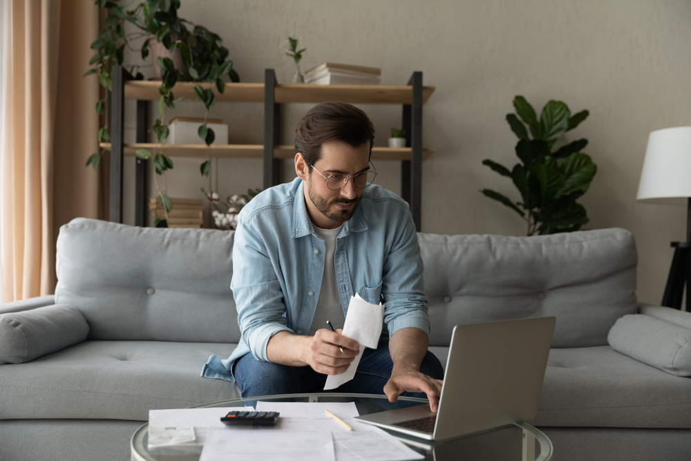 Man on couch looking at laptop and surrounded by paperwork.